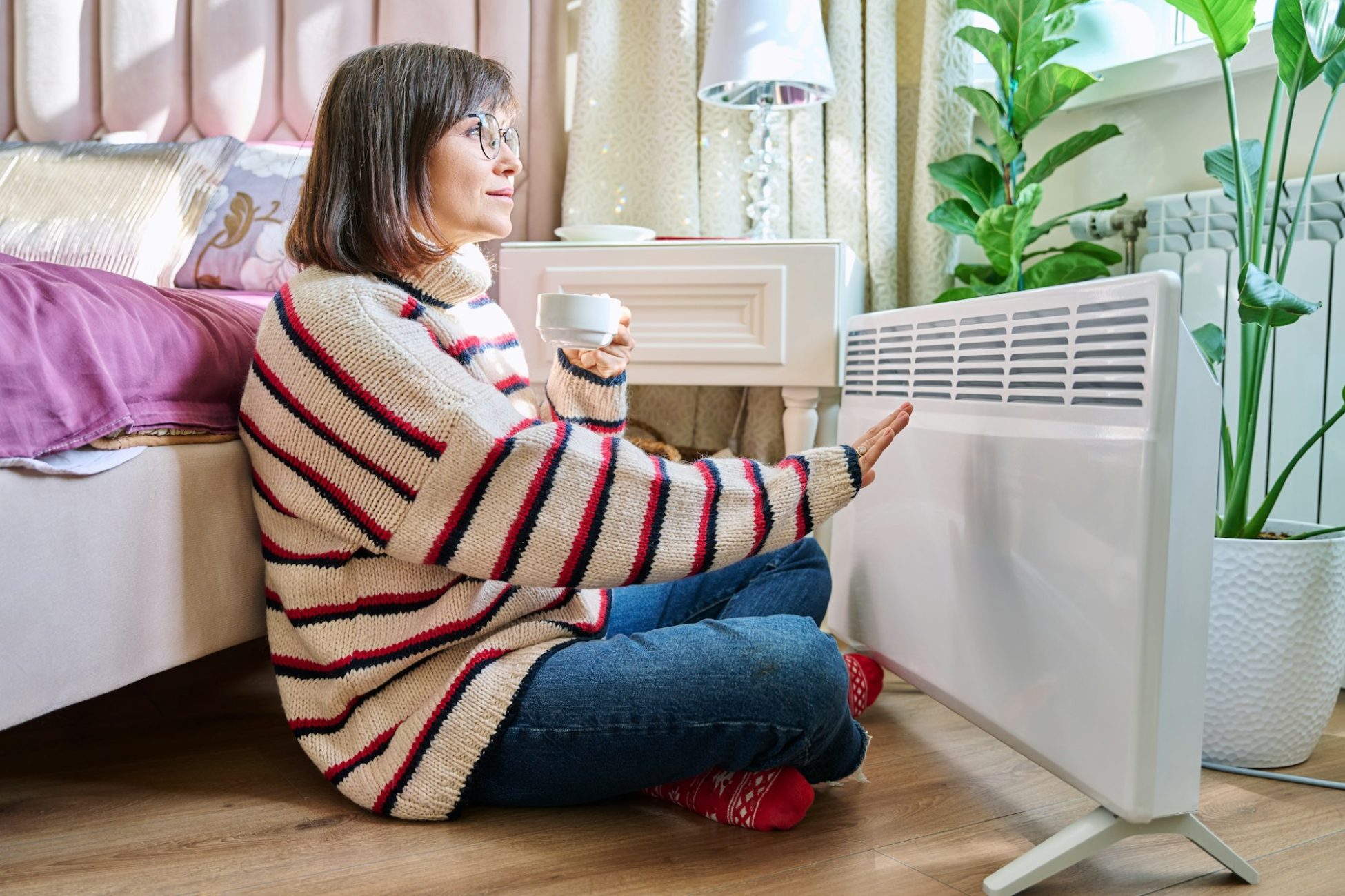 Woman warming herself in bedroom near electric heater